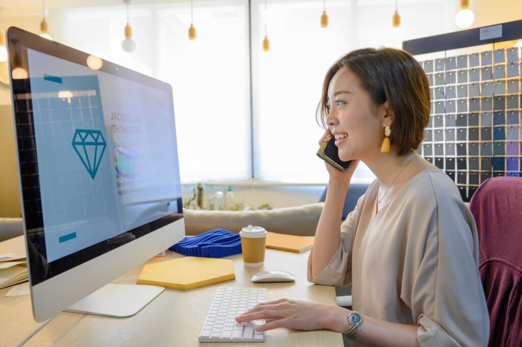 Woman at a desk talking on her phone while using a large computer monitor in a bright office, with a coffee cup nearby.