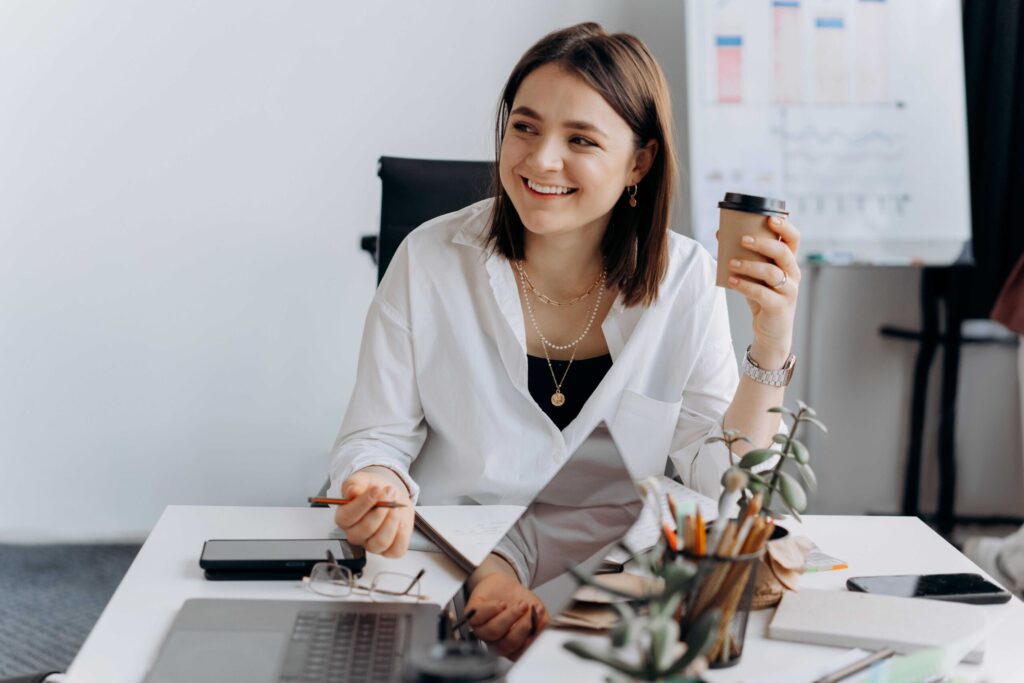 Smiling woman in a white shirt at a desk, holding a coffee cup and papers.