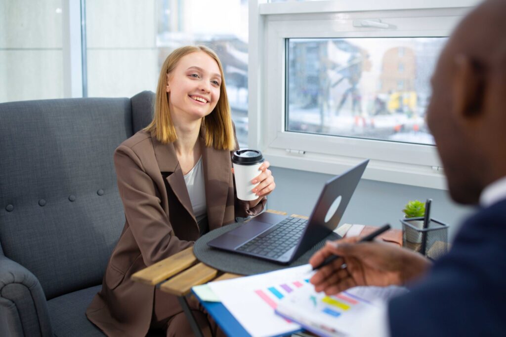 Smiling woman in a brown blazer sits with a coffee cup, laptop, and charts on the table during a discussion by a windowed office bench.