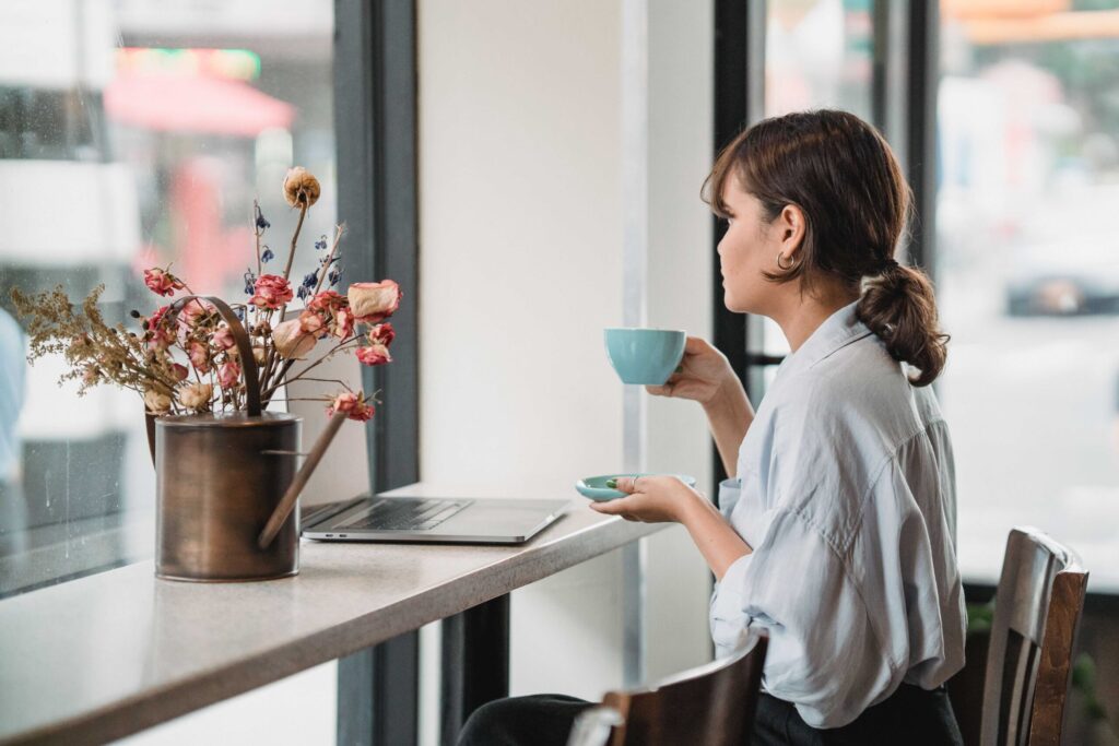 Woman seated at a cafe counter by a window, holding a blue teacup while looking at a laptop and flower vase nearby