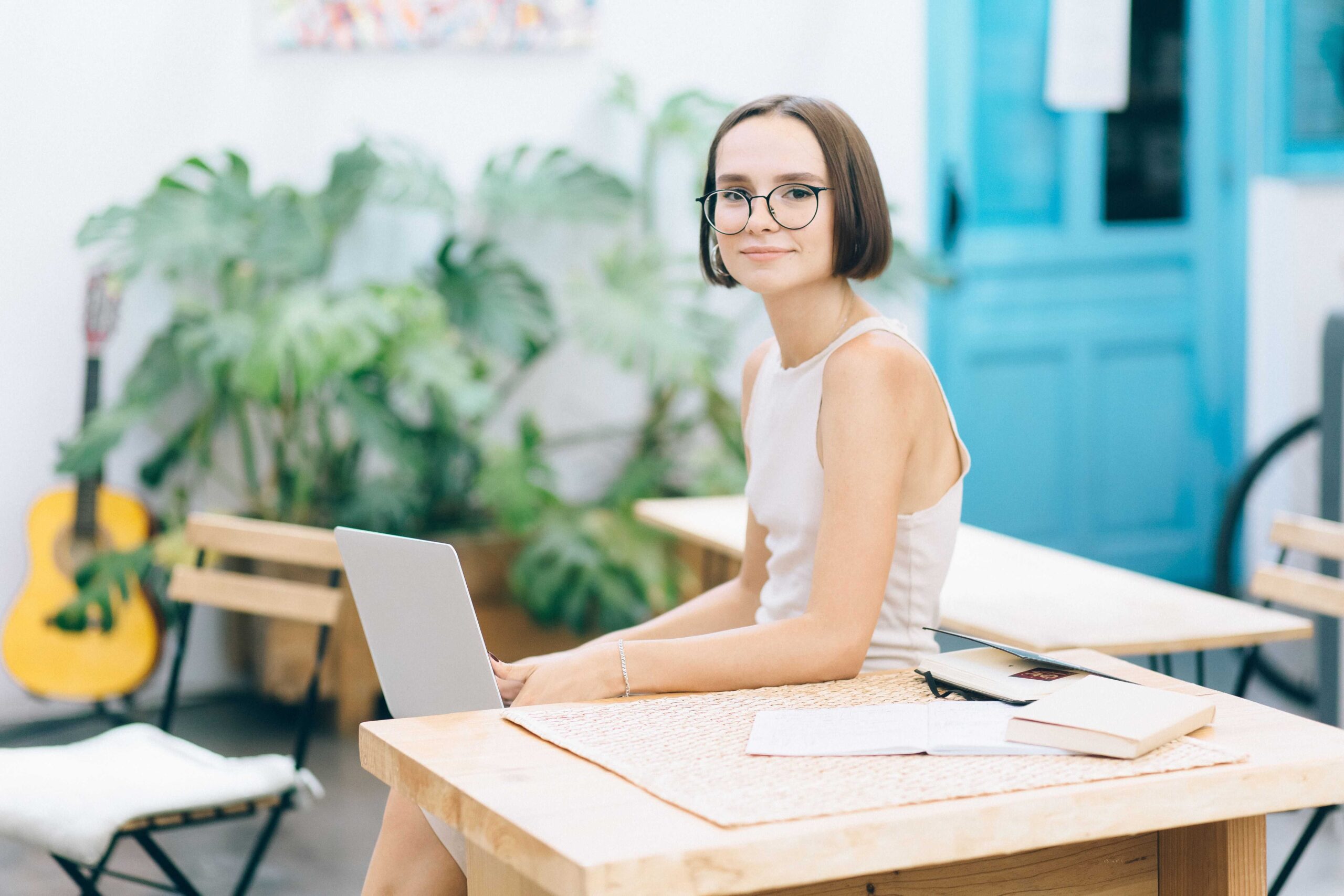 Woman with short brown hair and round glasses typing on a laptop at an outdoor cafe with greenery behind the table.