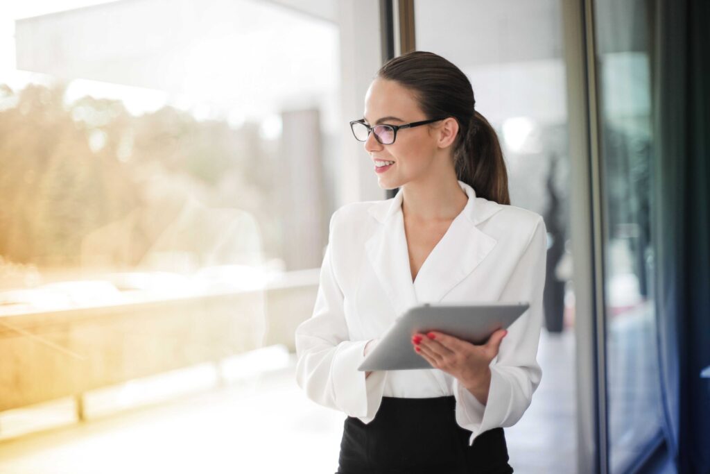 Professional woman in a white blazer holding a tablet, smiling as she looks out a glass wall