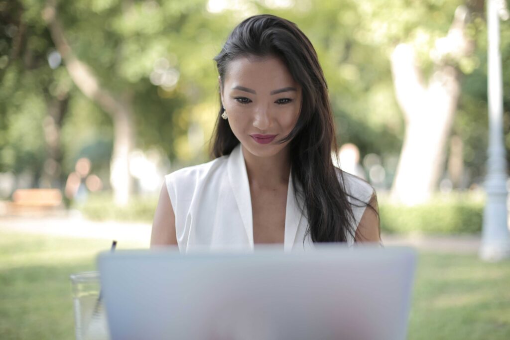 Woman with long dark hair in a white sleeveless top sits at an outdoor table, looking down at a laptop in front of her with a blurred green park background.