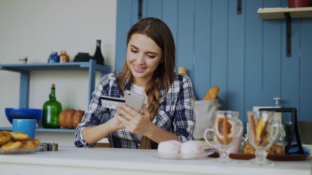 Young woman in a blue plaid shirt sits at a kitchen island, smiling while using her smartphone and a credit card in hand.