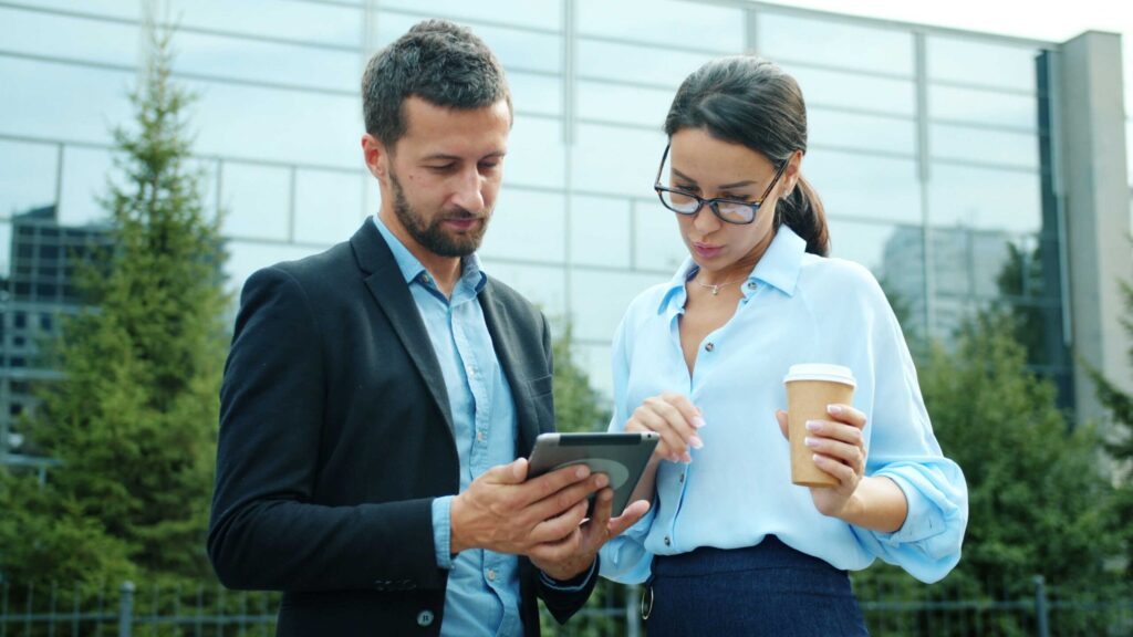 Two professionals outdoors, reviewing a tablet together, with a coffee cup in hand by one person.