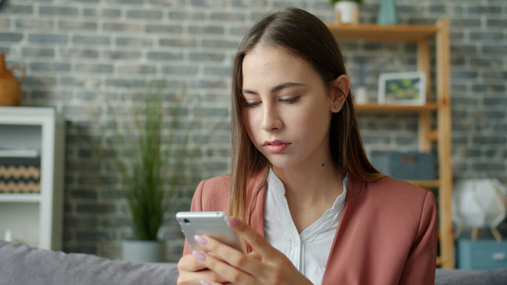 Woman in a pink blazer looking at her smartphone indoors in a living room setting.