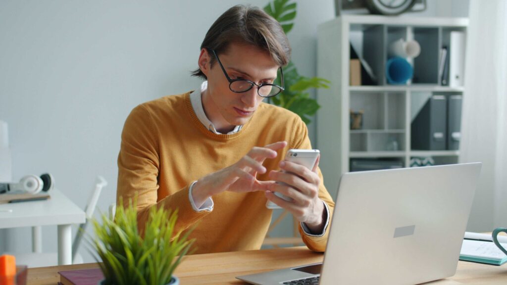 Person wearing glasses and a mustard sweater looks at a smartphone while seated at a desk with a laptop and plants nearby.