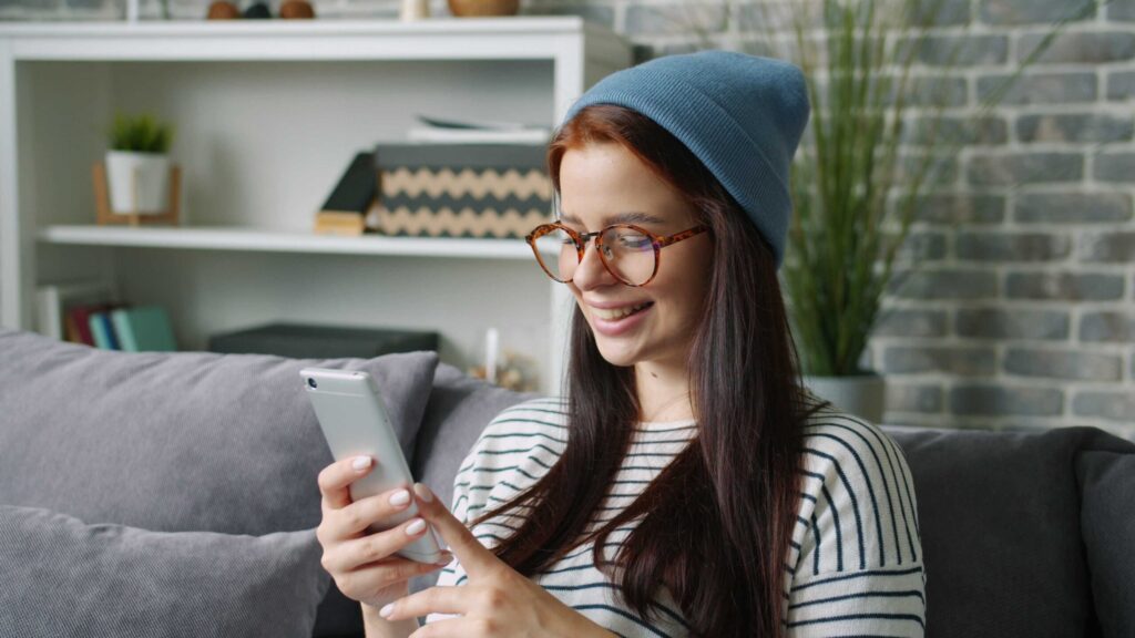 Woman wearing a blue beanie and tortoiseshell glasses sits on a gray couch, smiling while looking at her phone.