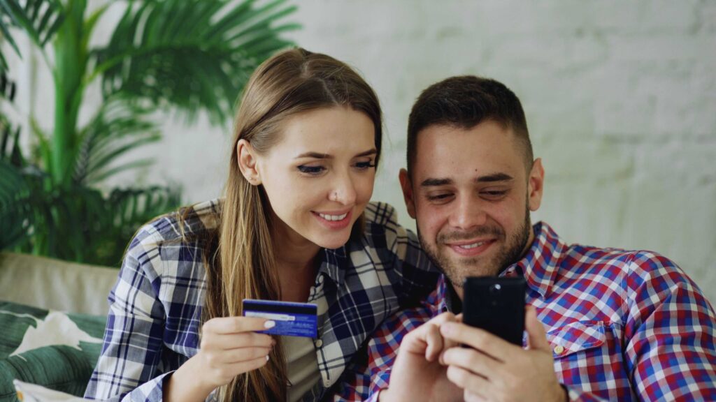 Young couple sitting on a sofa, looking at a smartphone; the woman holds a credit card as they shop online.