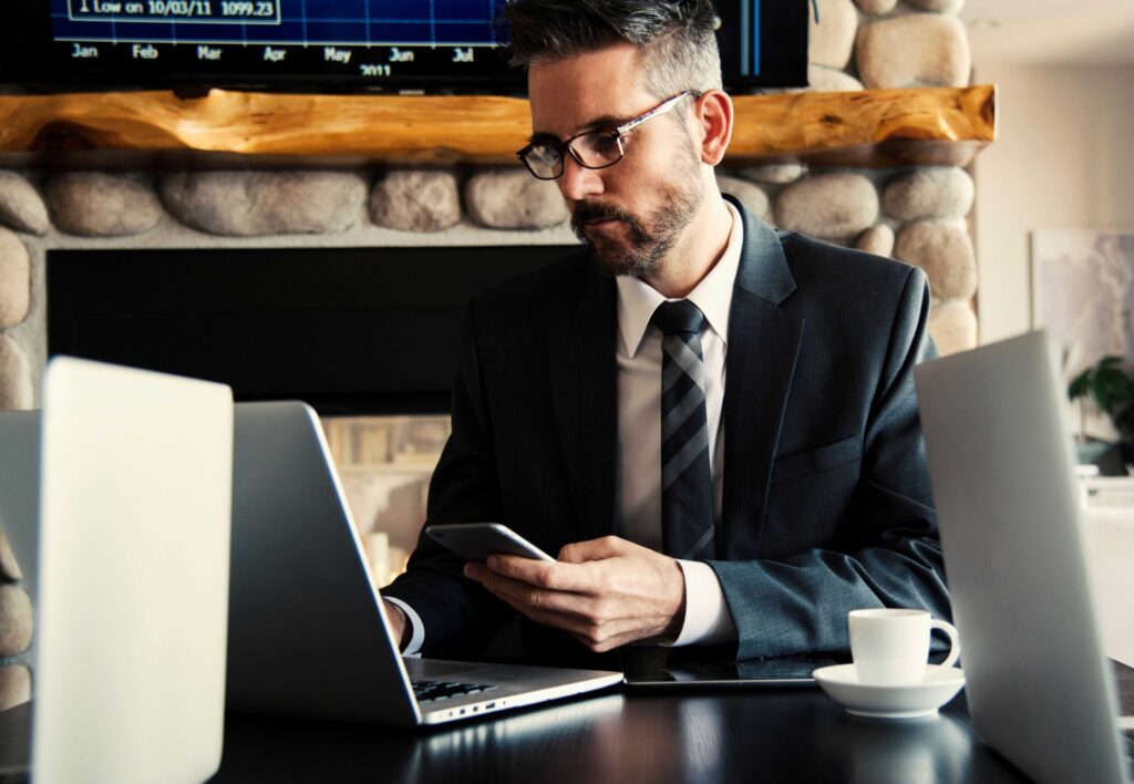 Man in a suit and glasses sits at a desk, looking at his phone with a laptop nearby in a modern workspace on a dark table.