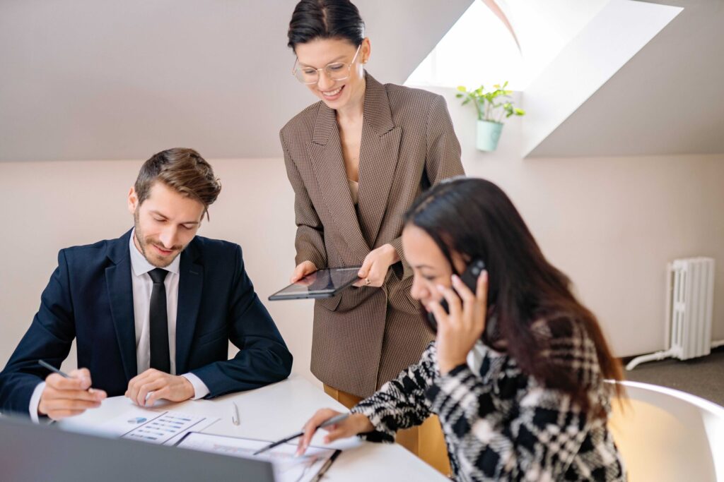 Colleagues in a bright office collaborate around a table; a man signs documents while a woman stands with a tablet and another woman talks on the phone.