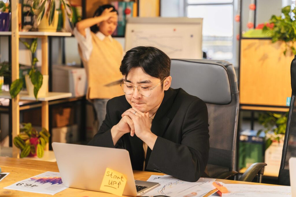 Businessperson wearing glasses sits at a laptop, hands pressed together, focused at a desk in a plant-filled office.