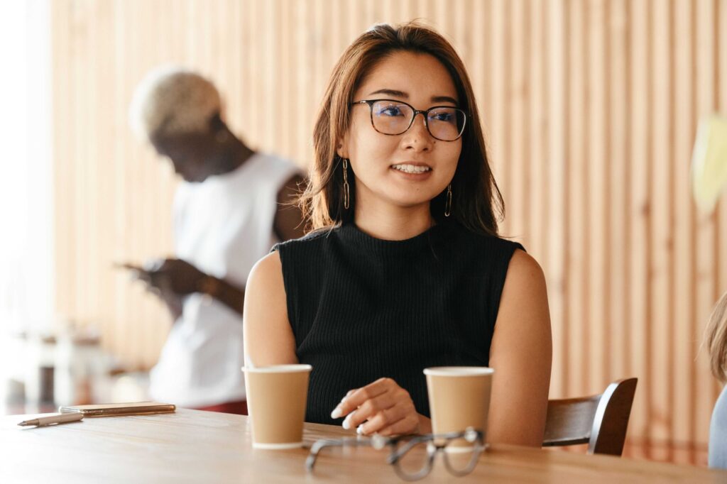 Woman with glasses seated at a cafe table, two coffee cups in front, blurred man in background behind her.