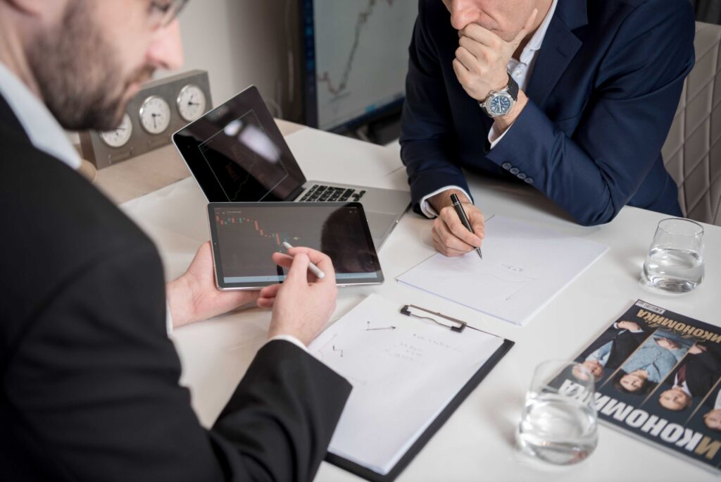 Two businessmen in suits discuss at a white-table conference, one showing a tablet with stock charts while the other writes notes.
