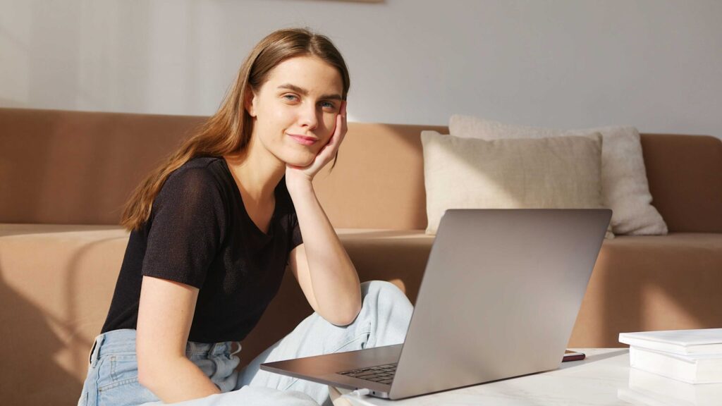 Young woman sitting on a beige sofa with a laptop, resting her head on her hand and smiling at the screen.