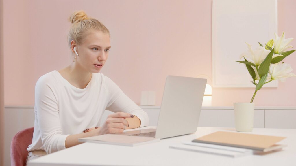Woman with blonde hair in a bun working at a white desk, wearing AirPods and a white top, with a laptop and a vase of white lilies nearby.