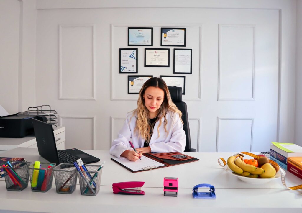 Woman seated at a white desk writing on paper in an office with framed certificates on the wall behind her and a laptop to her left.