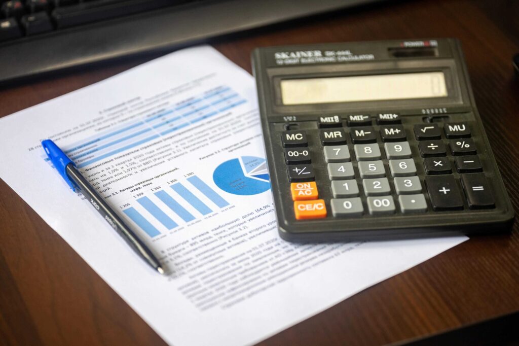 Calculator on a wooden desk beside printed financial reports with blue charts and a blue pen dipping into the page