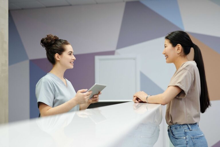 Two women at a reception counter: a nurse in light blue scrubs holds a tablet and smiles at a visitor in a beige top across the desk.