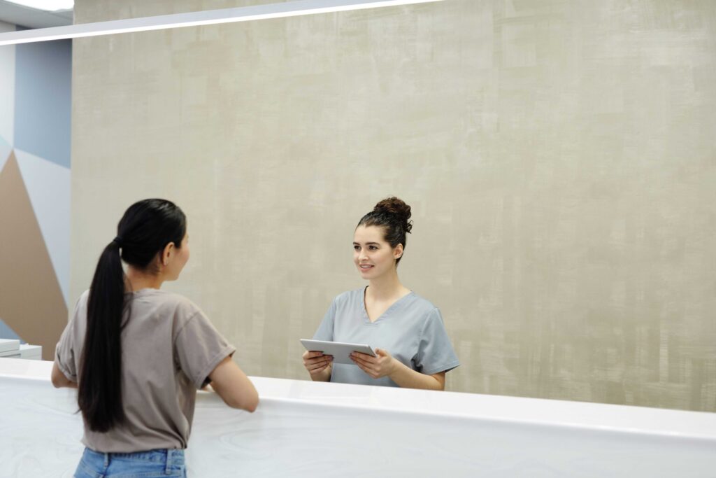 Reception nurse in gray scrubs holding a tablet speaks with a patient at a modern front desk in a clinic.