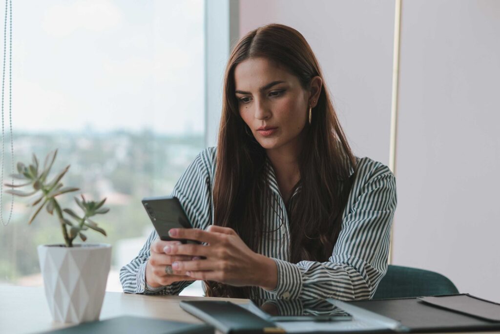 Woman in a striped shirt sits at a desk, looking down at her smartphone, with a potted plant nearby by the window in a bright office.