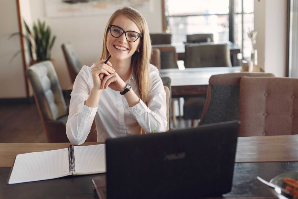 Smiling woman with glasses sits at a desk in a bright cafe, with a notebook and laptop in front of her.