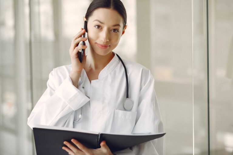 Female doctor in a white coat on a phone, holding a clipboard and stethoscope in a clinical setting