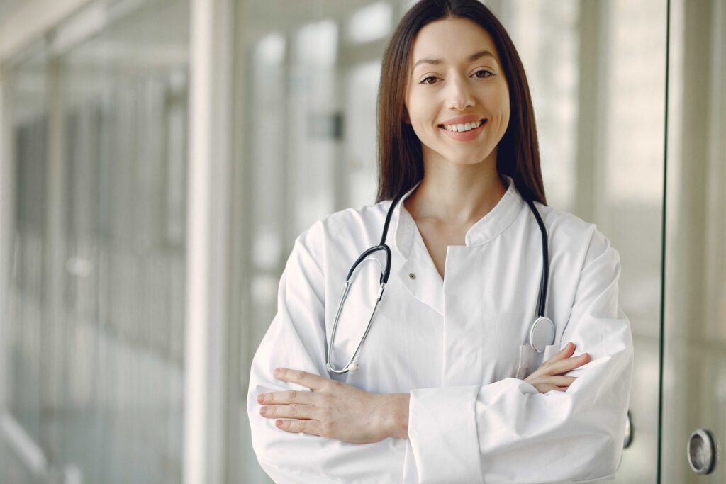 Smiling female doctor with stethoscope across white coat in a modern hallway, arms crossed confidently