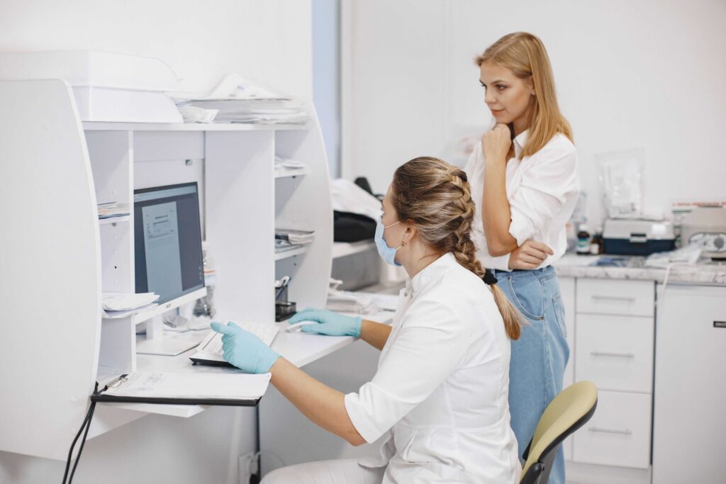 Two lab technicians in white coats work at a computer station; one seated wearing gloves and a mask, the other stands nearby looking at the monitor in a bright lab.