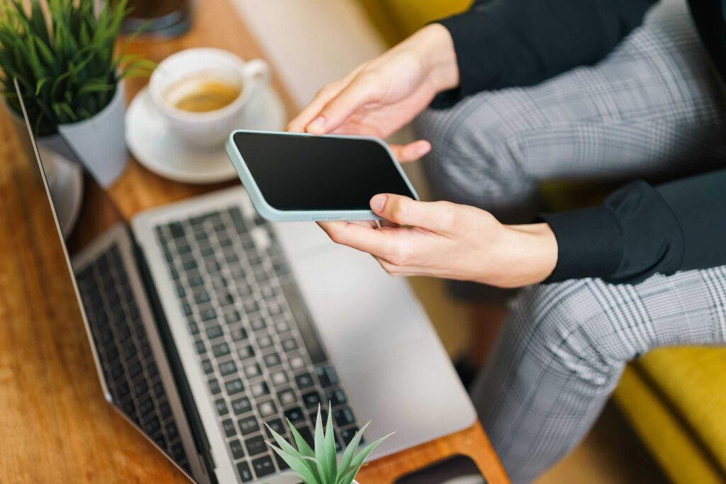 Person holding a smartphone over a laptop on a wooden desk, with a coffee cup and a small plant nearby.