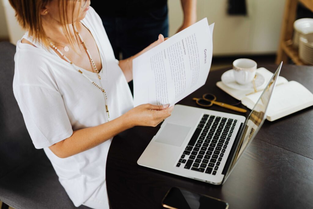 Person in a white blouse reads a printed document beside an open laptop on a dark wooden desk with a cup of coffee nearby.