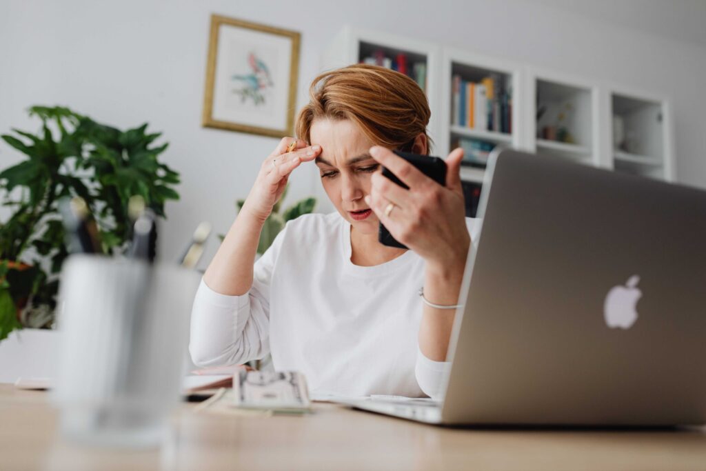 Woman at a desk holding a phone to her ear, frowning and touching her temple, in a home office with a plant and bookshelves in the background