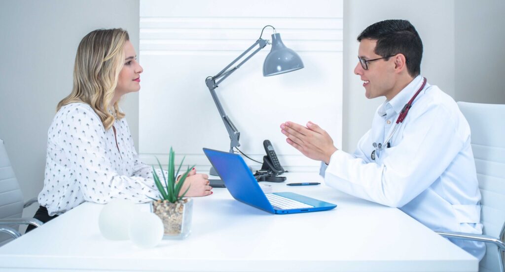 Doctor in white coat speaks with a female patient across a desk in a bright clinic, a blue laptop and phone on the table.