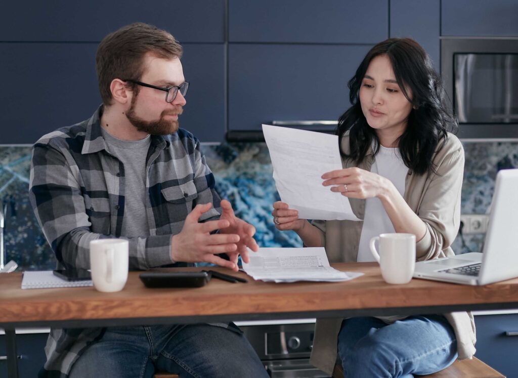 Two coworkers sit at a kitchen island, reviewing printed documents with a laptop and coffee mugs on the table, engaged in a discussion.