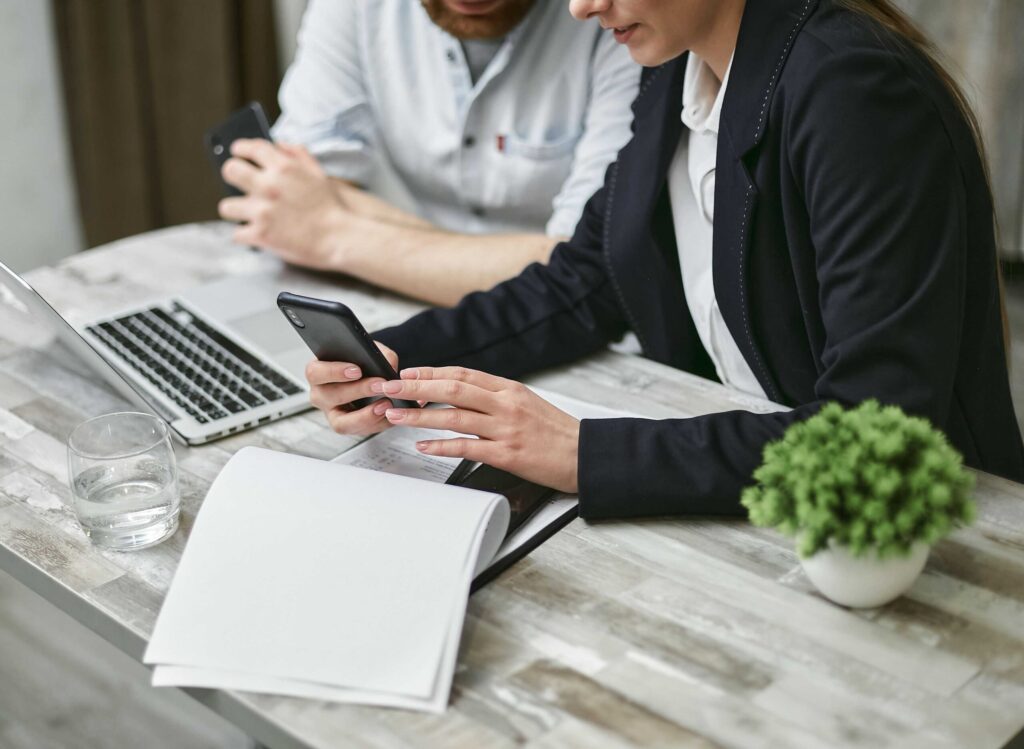 Two colleagues sit at a marble table, looking at a laptop and a smartphone amid papers and a small plant nearby.