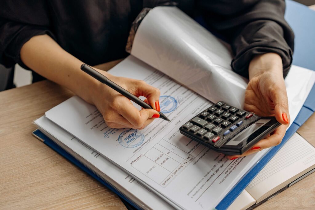 Hands using a calculator and writing on a stamped document on a desk, with a blue binder nearby.