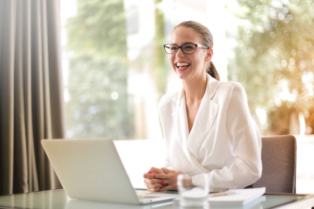 Smiling woman in a white blazer works on a laptop at a glass desk in a bright office.
