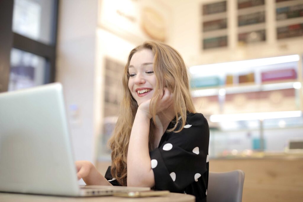 Woman with long hair smiling at a laptop in a bright cafe or study area, resting her chin on her hand.