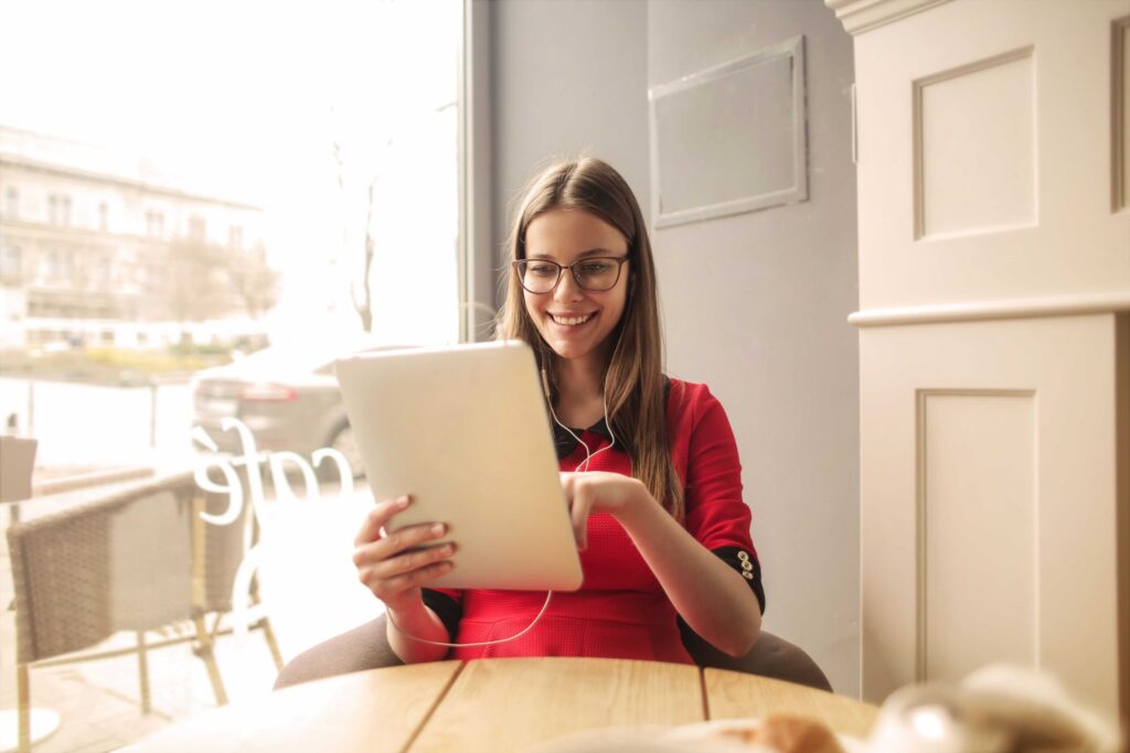 Smiling woman with glasses in a red dress, holding a tablet in a cafe by a window.