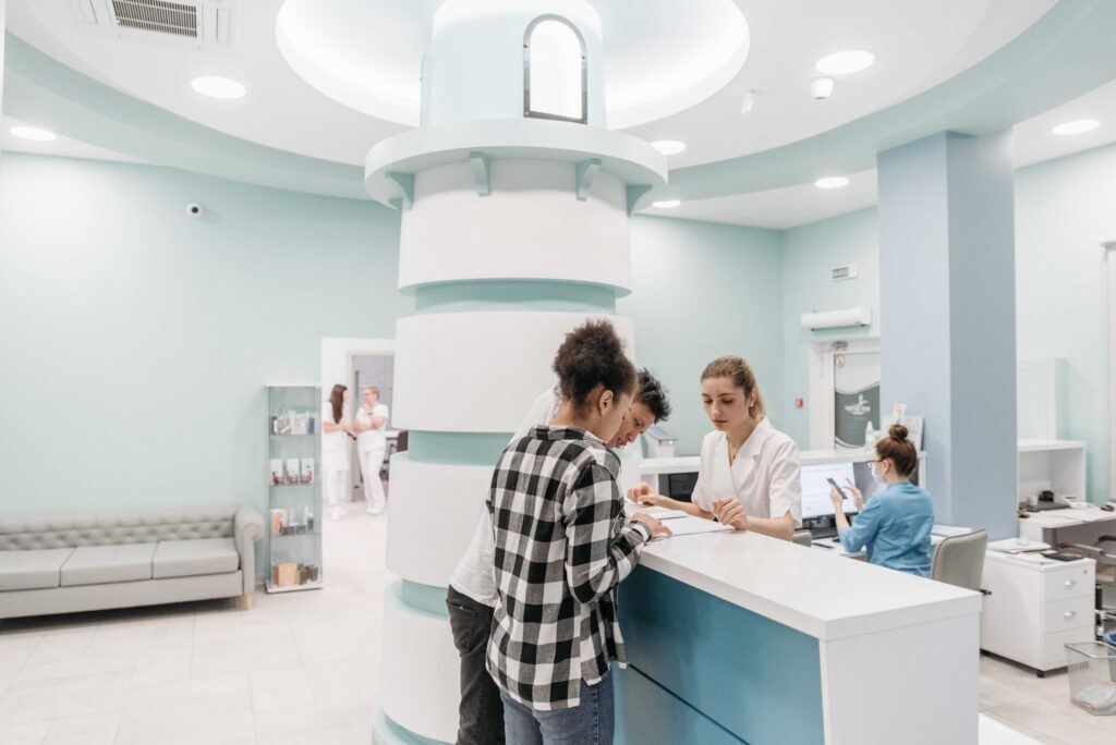 Medical reception area with staff assisting a patient at a curved teal-and-white counter in a bright clinic lobby