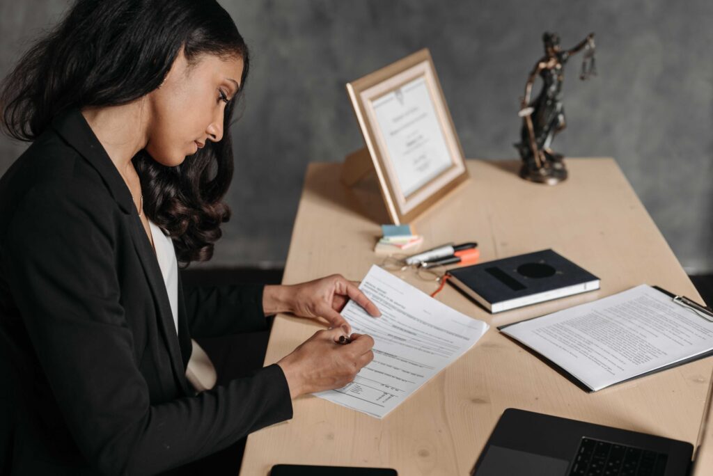 Professional woman in a blazer signs documents at a wooden desk with a framed certificate and statue in the background