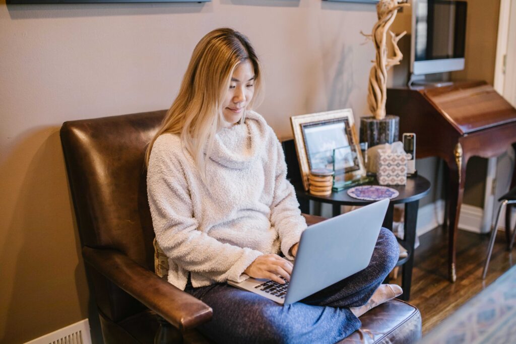 Woman in a cozy sweater sits on a leather chair using a laptop, indoors setting with framed photos on a side table behind her.