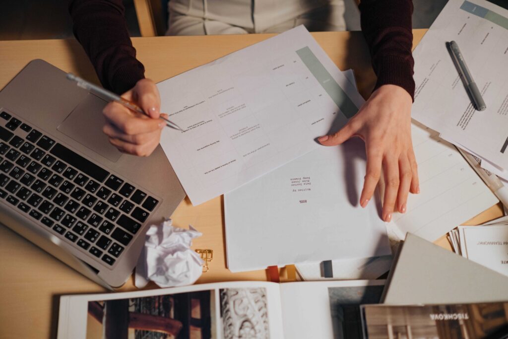 Overhead view of a person writing on a calendar page with papers and a laptop on a desk.