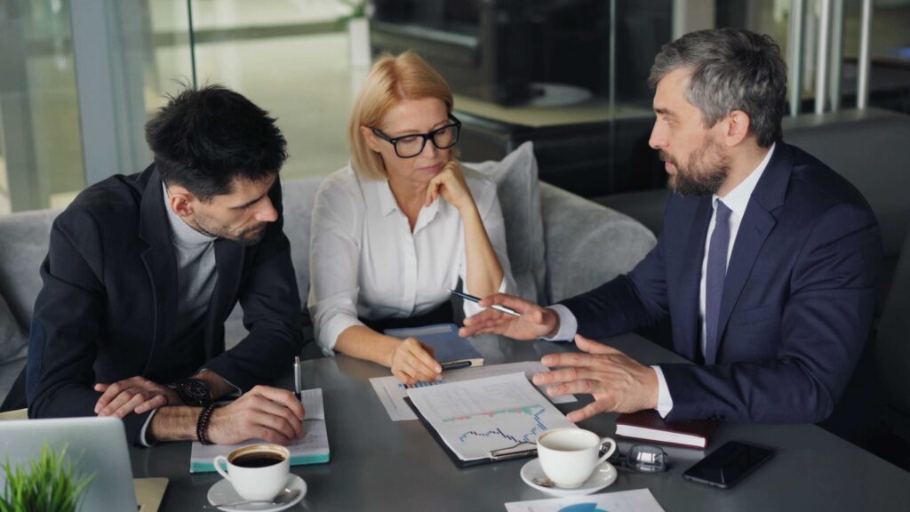 Three professionals in business attire sit at a table reviewing documents and charts during a meeting in a modern office setting, with coffee cups nearby.