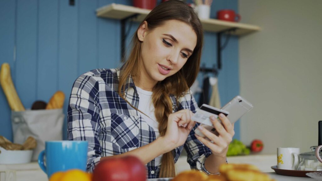 Woman in a blue plaid shirt holds a smartphone and card while standing in a kitchen with groceries on the counter.