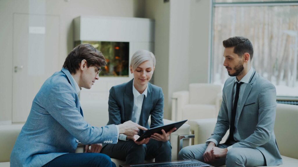 Three professionals in blue suits sit in a modern office, reviewing a tablet together during a meeting on a bright day.