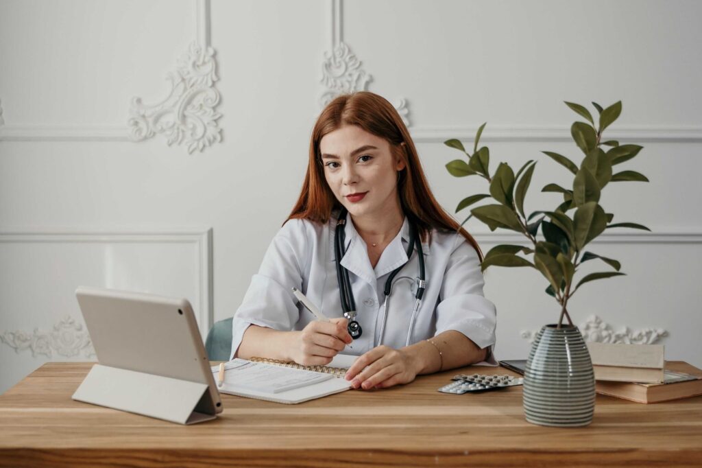 Female doctor in white coat with stethoscope sits at desk reviewing papers beside a laptop and a potted plant on a wooden table.