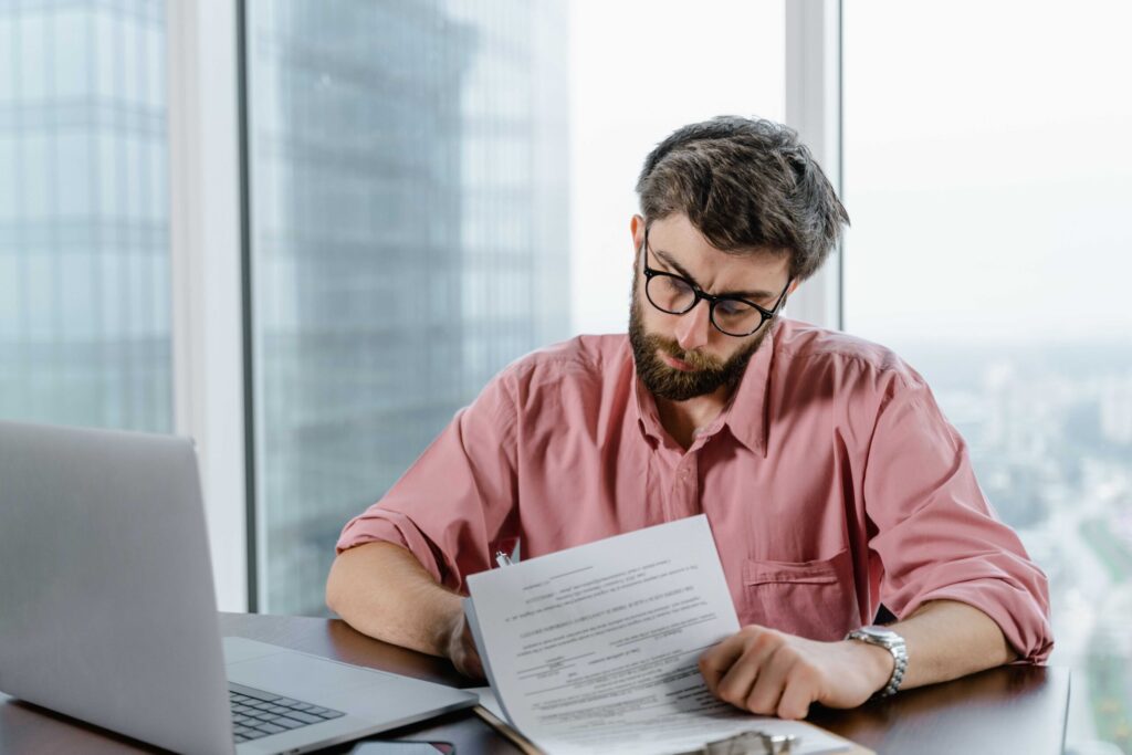 Bearded man in glasses wearing a pink shirt reviews a document at a desk beside a laptop in a bright office window.