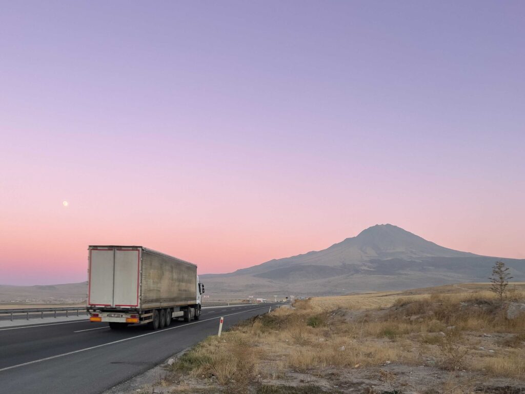 Semi truck on a highway at sunset with a distant mountain and pastel pink‑purple sky behind it.