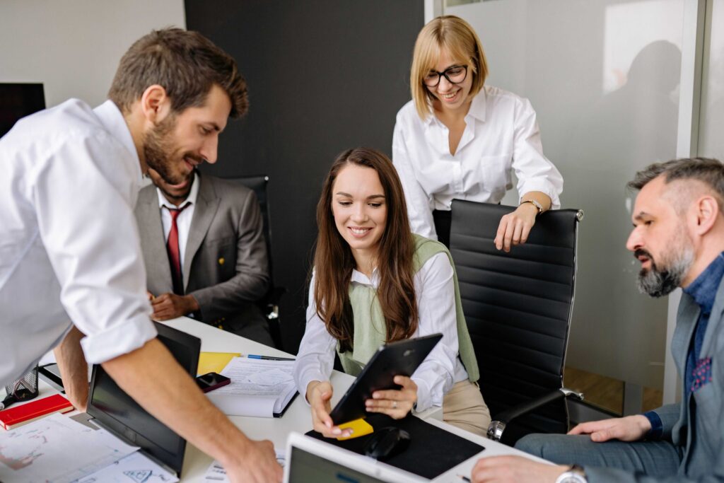 Five colleagues collaborate around a conference table with laptops, tablets, and papers in a bright office.